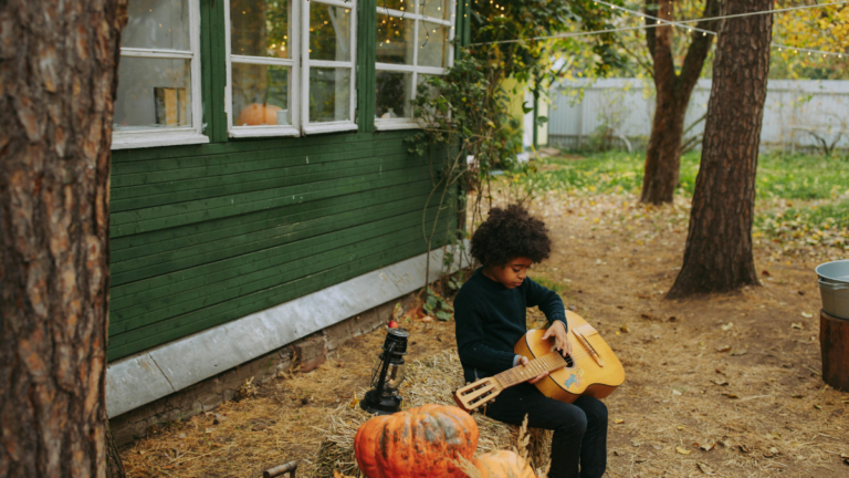 A child trying to play guitar near a pumpkin outside of his house